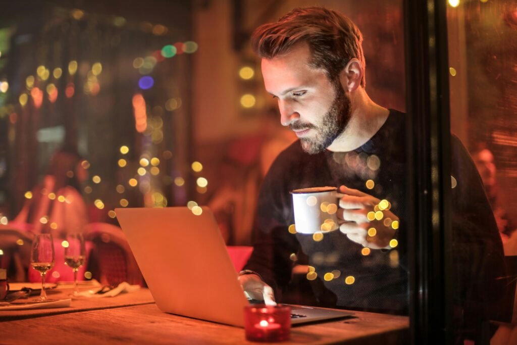 pexels-photo-842548-842548 A young man with a beard is working on a laptop at a cozy cafe with warm lighting, holding a coffee cup.