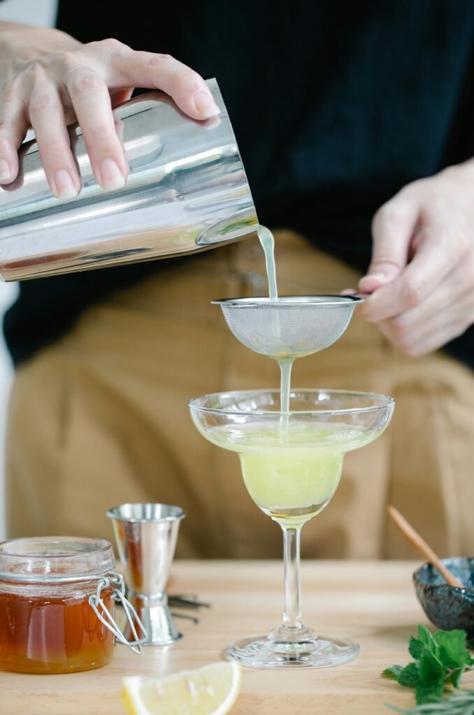 pexels-photo-8679149-8679149 A person pouring a freshly shaken cocktail through a sieve into a glass on a wooden table.